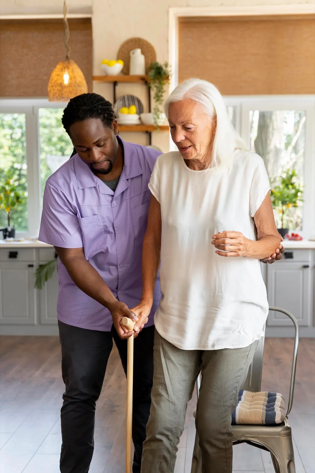 A carer walking beside an older woman with a stick in her kitchen